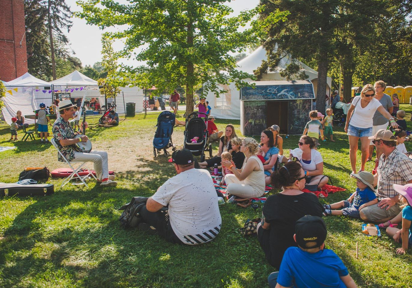 Families with young children sit on the grass, listening to a musician play a guitar